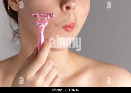 Una giovane donna con baffi tiene un rasoio alla guancia. Il concetto di sbarazzarsi dei capelli facciali indesiderati. Primo piano. Foto Stock