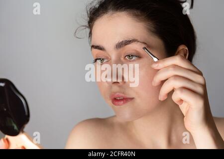 Giovane bella donna guarda in uno specchio compatto e corregge la forma delle sopracciglia. Il concetto di sbarazzarsi dei capelli indesiderati Foto Stock