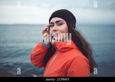 Ritratto di una donna brunetta dal look misterioso con capelli lunghi, con cappello e giacca arancione. Sullo sfondo il mare e la linea dell'orizzonte Foto Stock