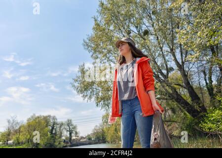 Donna volontaria in piedi sulla riva del fiume con un sacco di rifiuti. Il concetto di inquinamento ambientale. Cielo e alberi sullo sfondo. Foto Stock