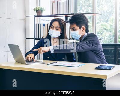 Uomo d'affari asiatico e donna che indossano abiti e maschere facciali di protezione utilizzando il computer sulla scrivania, riunioni e lavoro insieme in ufficio, nuovo lavoro normale Foto Stock