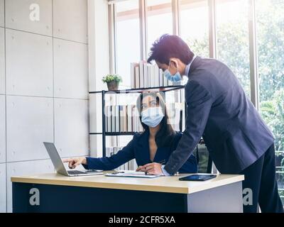 Uomo d'affari asiatico e donna che indossano abiti e maschere facciali di protezione utilizzando il computer sulla scrivania, riunioni e lavoro insieme in ufficio, nuovo lavoro normale Foto Stock