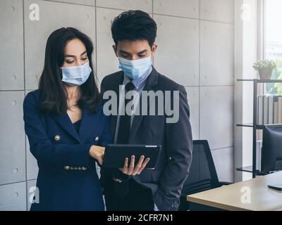 Uomo d'affari asiatico e donna che indossano abiti e maschere facciali di protezione utilizzando tablet digitale, riunioni e lavoro insieme in ufficio, nuovo lavoro normale l Foto Stock