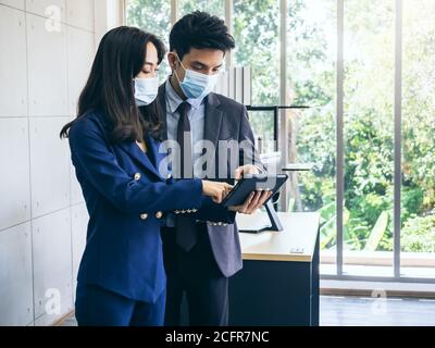 Uomo d'affari asiatico e donna che indossano abiti e maschere facciali di protezione utilizzando tablet digitale, riunioni e lavoro insieme in ufficio, nuovo lavoro normale l Foto Stock