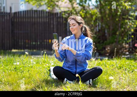 Una bella donna felice è seduta a gambe incrociate sull'erba, utilizzando un telefono cellulare. Sullo sfondo c'è una recinzione. Giornata di sole, all'aperto. Concetto di calza Foto Stock