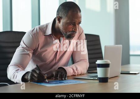 Uomo d'affari senior prende appunti mentre guarda il suo laptop a. ufficio moderno Foto Stock