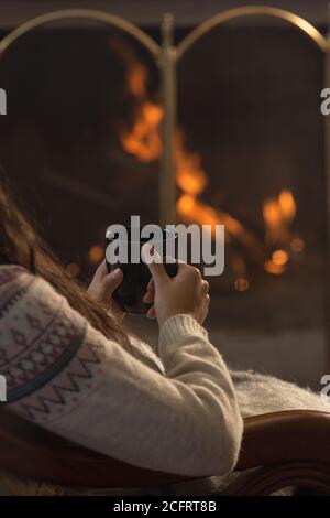 persona con un maglione di lana e i capelli lunghi seduti dentro davanti ad un camino illuminato e tenendo una tazza nera nelle sue mani Foto Stock