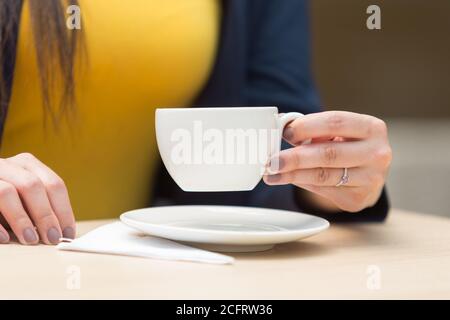 mano con anelli dipinti con un anello che tiene e solleva una piccola tazza bianca, c'è un piatto e un tovagliolo, su una superficie liscia di legno, sullo sfondo Foto Stock