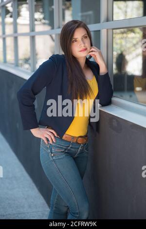 Donna con pelle bianca latina e capelli lunghi, in posa in piedi accanto a una finestra, indossa una blusa gialla, una giacca blu, jeans e cintura, foto di giorno Foto Stock