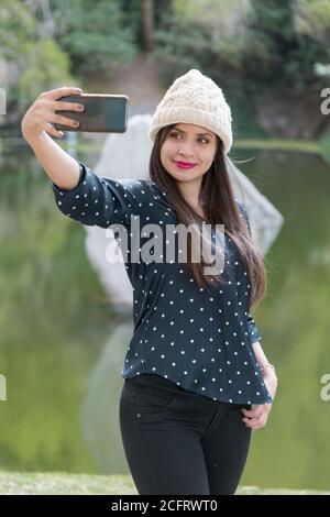 Giovane donna dalla pelle bianca con trucco e capelli lisci, scattando una foto con il suo cellulare, indossando un cappuccio di lana e una blusa nera, selfie Foto Stock