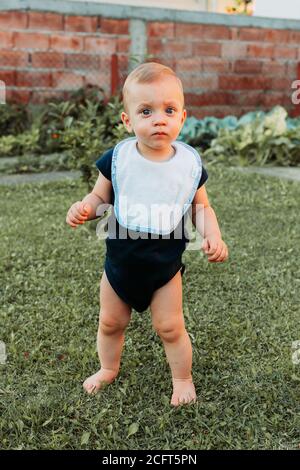 L'adorabile ragazzo di un anno si trova nell'erba a piedi nudi, mantenendo il suo equilibrio, sembra confuso Foto Stock
