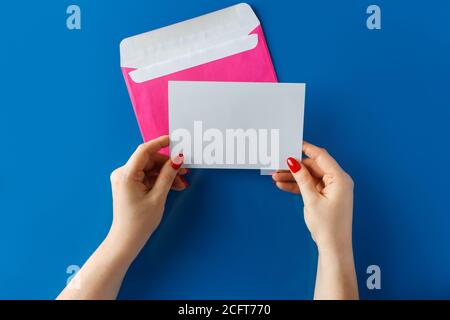 Busta rosa con una scheda vuota in mani su sfondo blu. Mani che tengono una busta rosa con una lettera vuota su sfondo blu. Foto Stock