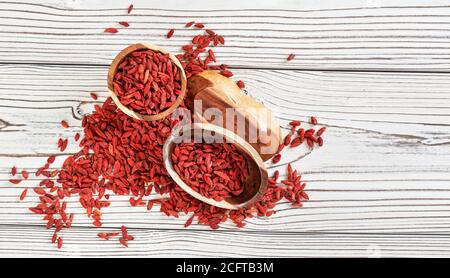 Bacche di goji secche in ciotole di legno, sparse su tavole bianche sotto, vista dall'alto Foto Stock