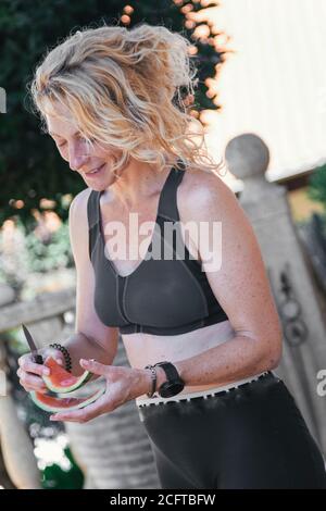 Giovane donna bionda matura in abbigliamento sportivo mangiare melone all'aperto Foto Stock