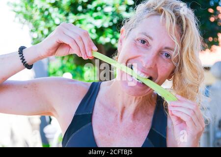 Giovane bionda matura in abbigliamento sportivo che batte un gambo di sedano all'aperto Foto Stock