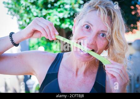 Giovane bionda matura in abbigliamento sportivo che batte un gambo di sedano all'aperto Foto Stock