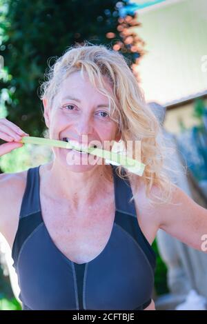 Giovane bionda matura in abbigliamento sportivo che batte un gambo di sedano all'aperto Foto Stock