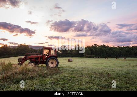 Il trattore rosso e le balle di fieno appena rotolate poggiano sul rotolamento collina con suggestivo paesaggio nuvoloso all'alba, creando un rustico rurale scena Foto Stock