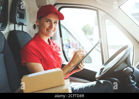 giovane sorridente delivery in uniforme rossa seduta in furgone e la scrittura di documenti Foto Stock
