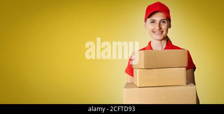 servizio di consegna - giovane donna sorridente in rosso uniforme tenuta scatole di cartone davanti su sfondo giallo con spazio per la copia Foto Stock