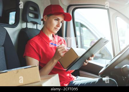 donna di consegna in uniforme rossa seduta in furgone e scrittura documenti Foto Stock