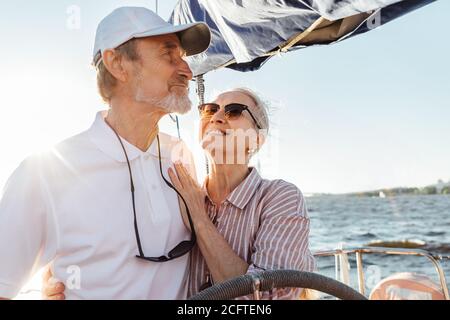 Felice donna matura che guarda il marito mentre guida uno yacht. Una coppia affettuosa che si gode una vacanza in barca a vela privata in una giornata di sole. Foto Stock