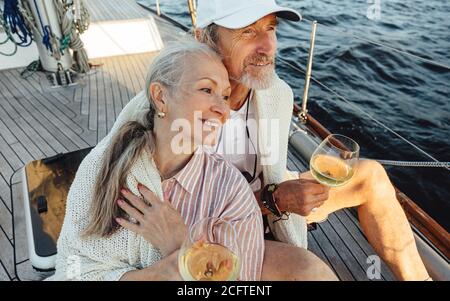 Coppia senior avvolta con plaid e seduta su un ponte di yacht con bocali di vino. Due persone affettuose e mature che si siedono insieme su una barca a vela e che guardano Foto Stock