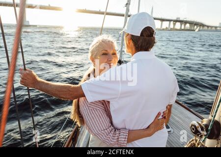 Sorridente femmina guarda sopra la spalla del marito e lo abbracciava. Coppia affettuosa in piedi insieme sulla barca a vela. Foto Stock