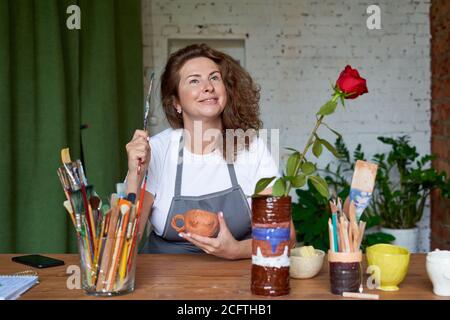 Felice imprenditore che si siede nel negozio di lavoro e sorridente. Donna artigiana che ha una buona idea. L'artigiano creativo è felice e fiducioso. L'artista è seduto a tavola e sogna Foto Stock