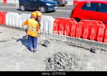 I lavoratori della strada riparano un vecchio tratto di strada con un martello pneumatico e una pala, rimuovendo il vecchio calcestruzzo usurato dalla strada. Il passaggio delle auto in una sfocatura, Foto Stock