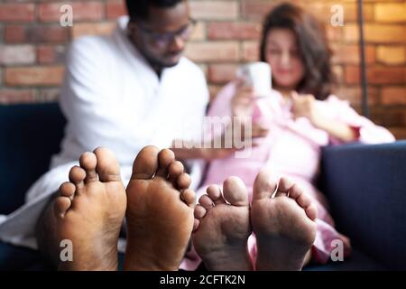 primo piano foto di piedi, coppia interracial hanno riposo a casa, aspettandosi bambino, parlare seduto sul divano Foto Stock