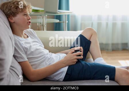 giocoso adolescente appassionato di videogiochi, sedersi sul divano in soggiorno tenendo la console in mano, godere, tempo libero Foto Stock