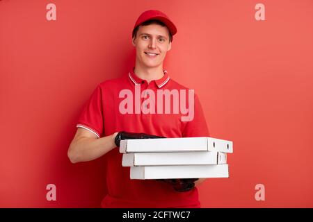 uomo in uniforme rossa con ordine di fast food isolato su sfondo rosso, corriere maschio che tiene la pizza per i clienti, indossare guanti protettivi Foto Stock