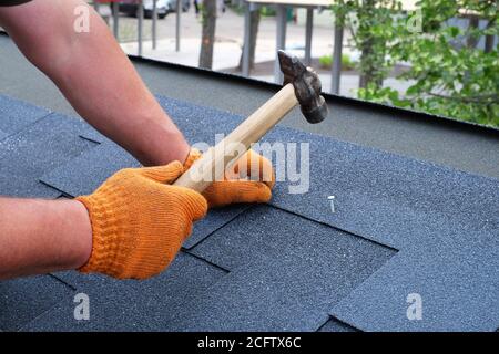 Mani di lavoro che installano gli shingle del tetto di bitume usando il martello nei chiodi. Foto Stock