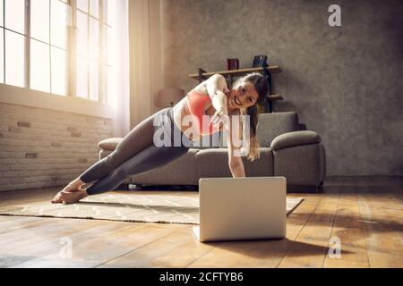 La ragazza giovane segue con un laptop un esercizi di palestra. È a casa a causa della quarantena di coronavirus codiv-19 Foto Stock
