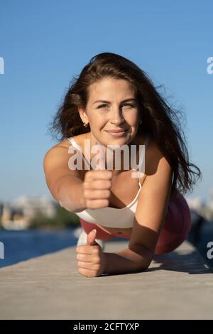 Fit gioiosa donna in rosa leging facendo esercizio in tavola lavorando su muscoli addominali e triceps, mostrando pollice in su, all'aperto. Sport modello femminile facendo c Foto Stock