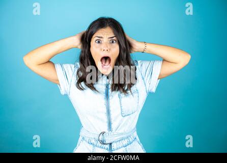 Giovane bella donna che indossa un jumpsuit in denim su sfondo blu isolato sorpreso e spaventato con le mani in testa. Foto Stock