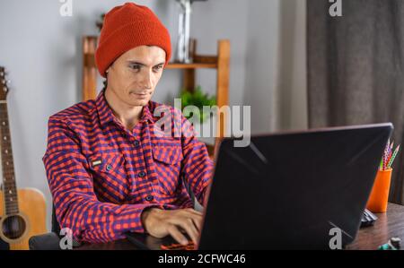 Un giovane moderno in una camicia rossa controllata e cappello sta lavorando su un computer portatile. Concetto di comunicazione a distanza e lavoro remoto. Foto Stock