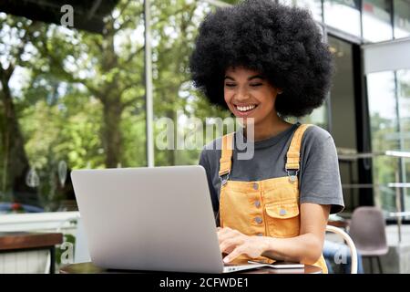 Felice studentessa africana che usa un computer portatile seduto al tavolo in un caffè all'aperto. Foto Stock