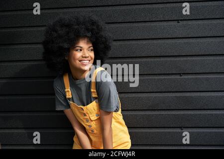 Donna africana di stile felice con i capelli afro che guardano via levano in piedi contro parete nera. Foto Stock