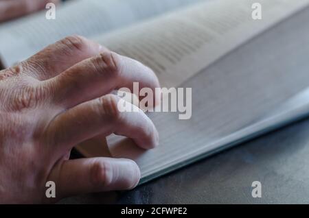 Mano maschile matura dall'alto su un libro aperto. Copertina rigida spessa. Mano maschile con un libro su un tavolo nero. Messa a fuoco selettiva. Foto Stock