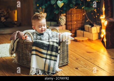 Carino bambino con il suo giocattolo preferito in un cesto di vimini sul pavimento vicino all'albero di Natale e regali. Stagione di festa. Vacanze di Capodanno. Foto Stock