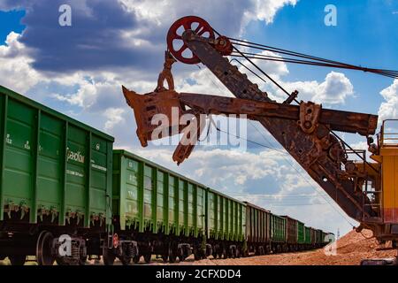 Estrazione e trasporto di minerali di alluminio. Attività minerarie aperte. Caricamento di argilla bauxite con escavatore elettrico nel treno tramogge. Foto Stock