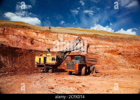 Argilla bauxite aperta taglio miniera. Estrazione e trasporto di minerali di alluminio. Escavatore giallo carico dumper da cava arancione Hitachi. Su suolo rosso e cielo blu Foto Stock