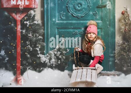 Carina bambina seduta vicino alla porta della casa la sera di Natale. Bambino cammina per la strada con una slitta da sola Foto Stock