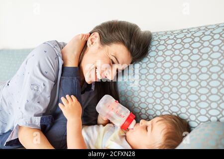 La madre felice dà il biberon del latte al suo bambino sul divano nel soggiorno Foto Stock