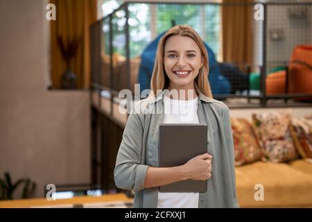 Ritratto di una giovane allegra donna caucasica, lavoratrice d'ufficio che tiene un tablet digitale e guarda la fotocamera mentre si trova in un ufficio moderno Foto Stock