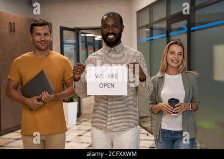 Giovane felice lavoratore africano ufficio mostrando carta con testo NOI SONO APERTI alla fotocamera e sorridono mentre si sta in piedi con la sua colleghi nell'ufficio moderno Foto Stock