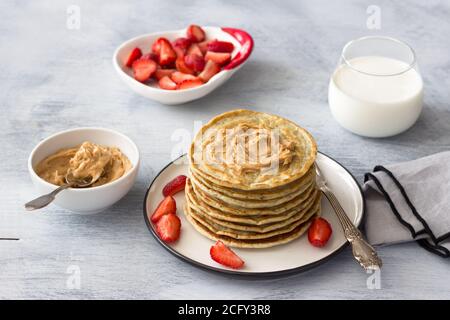 Frittelle vegane con semi di lino, burro di arachidi, fragola e latte vegano su un piatto bianco su sfondo grigio. Deliziosi cibi sani fatti in casa Foto Stock