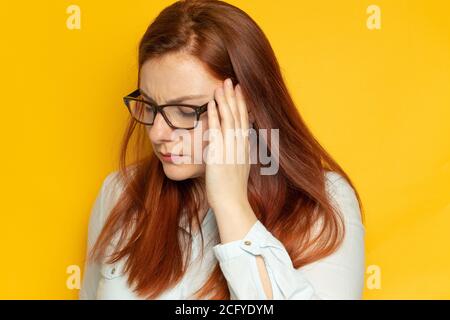 Donna giovane operaia in vista occhiali e con mal di testa su sfondo giallo parete. Concetto di stress, fatica. Le mani tengono la testa, cercando di trovare una soluzione al loro problema Foto Stock
